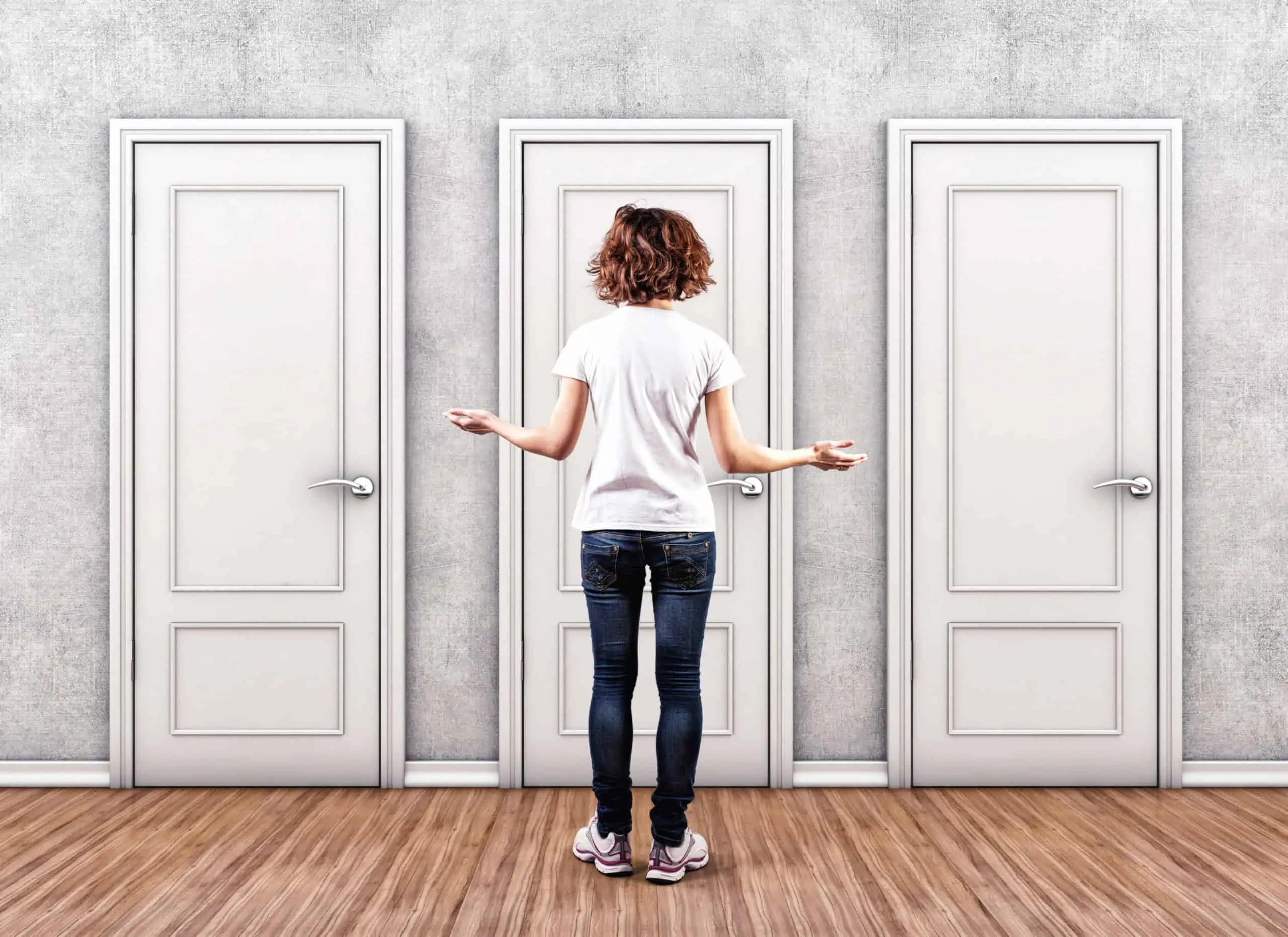 A redheaded Caucasian woman in dark blue denim jeans and a white t-shirt standing in front of multiple doors deciding how to file taxes.