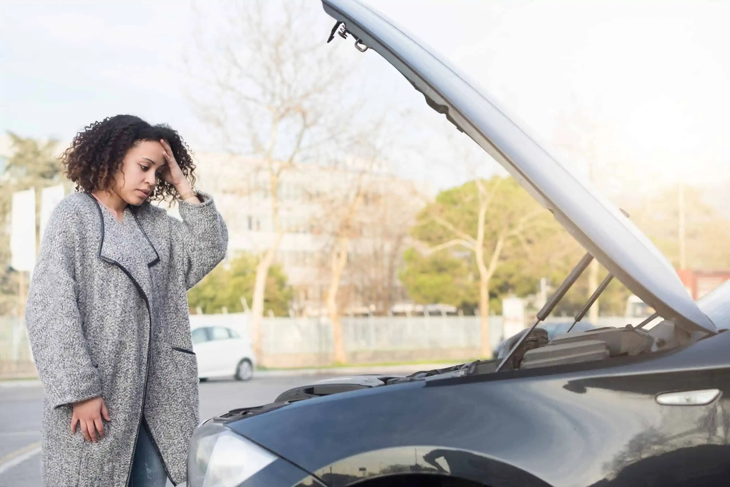 Black woman with curly hair, wearing grey overcoat looking in despair at car in need of repair