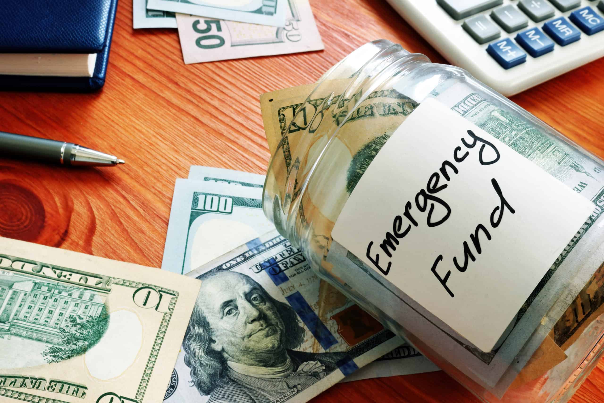 Glass jar labeled "emergency fund" with ten-dollar bills and hundred dollar bills on a Cherrywood desk