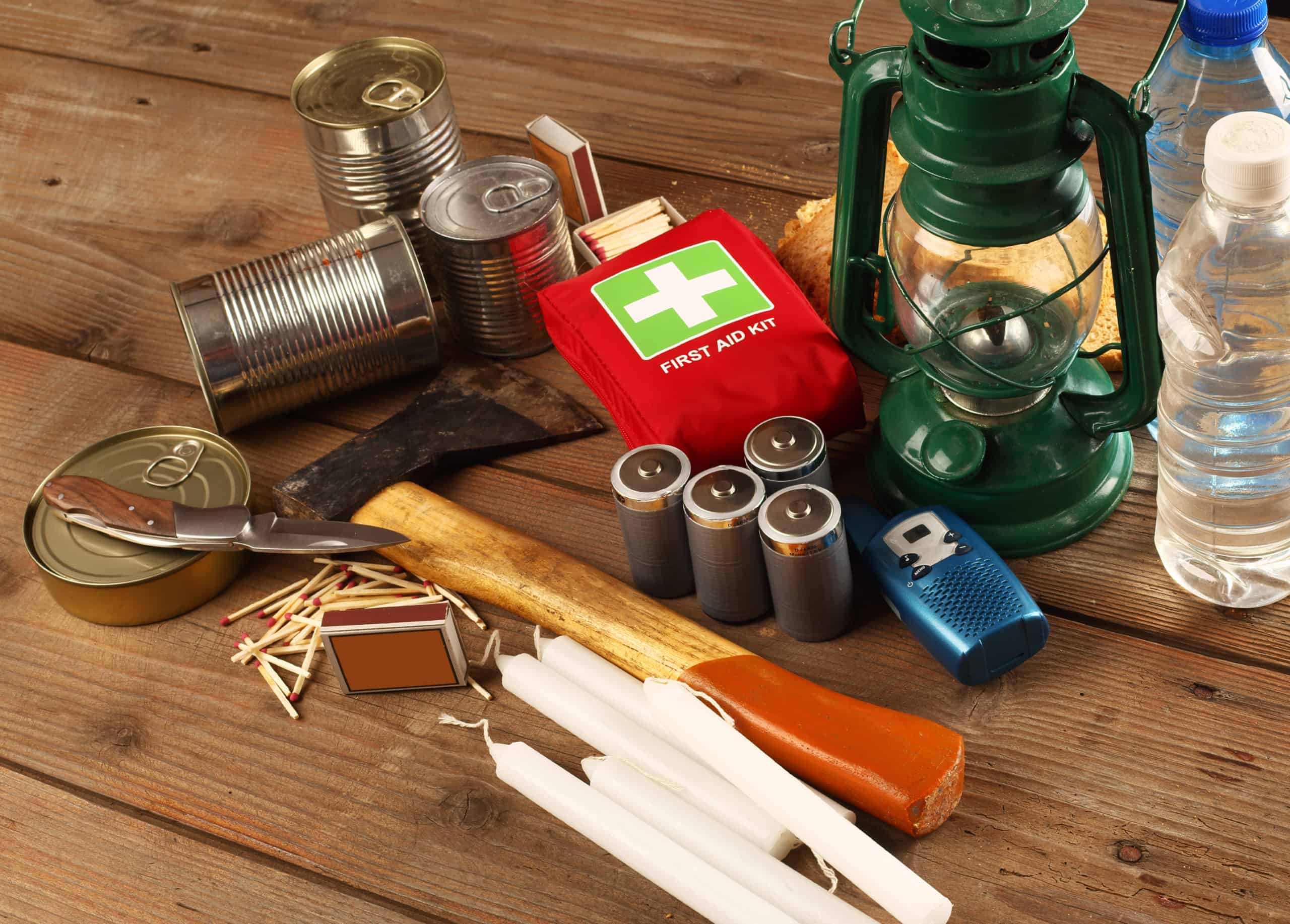 Emergency preparedness supply kit including an oil lantern, first aid kit, batteries, candles, matches, canned food, bottled water, knife, hatchet on a wooden table