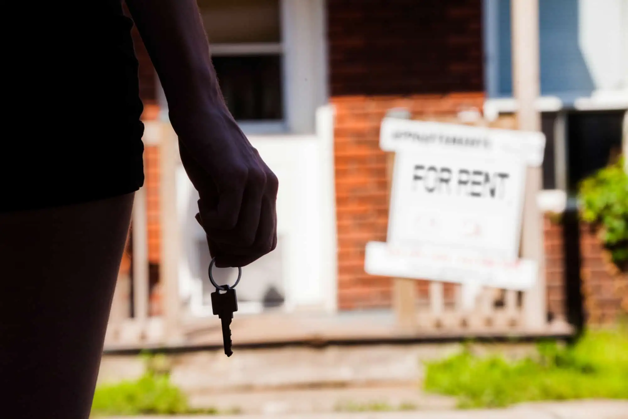 Caucasian woman holding keys to a house standing in front of a "for rent" sign in the front yard.