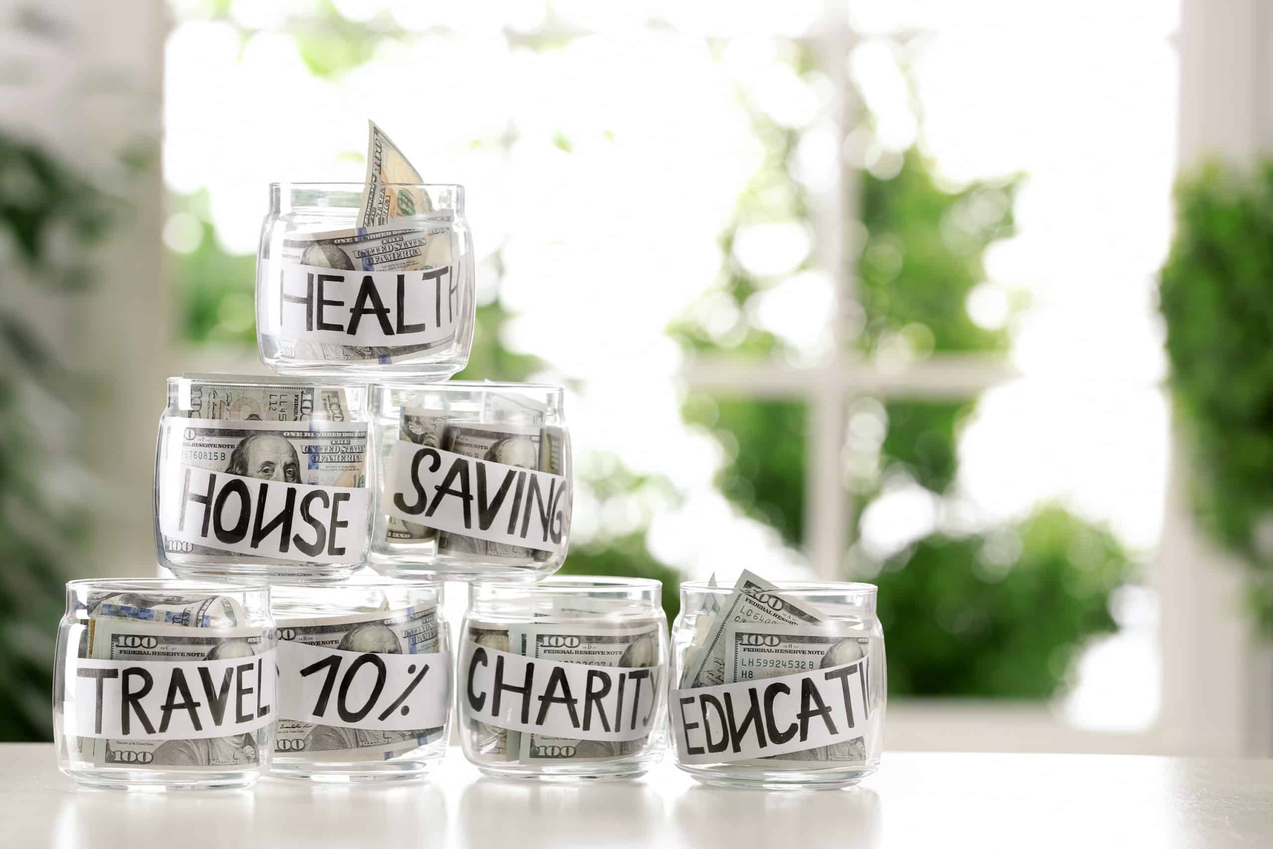 $100 bills stuffed into glass jars labeled health, house, saving, travel, charity and education for money management purposes