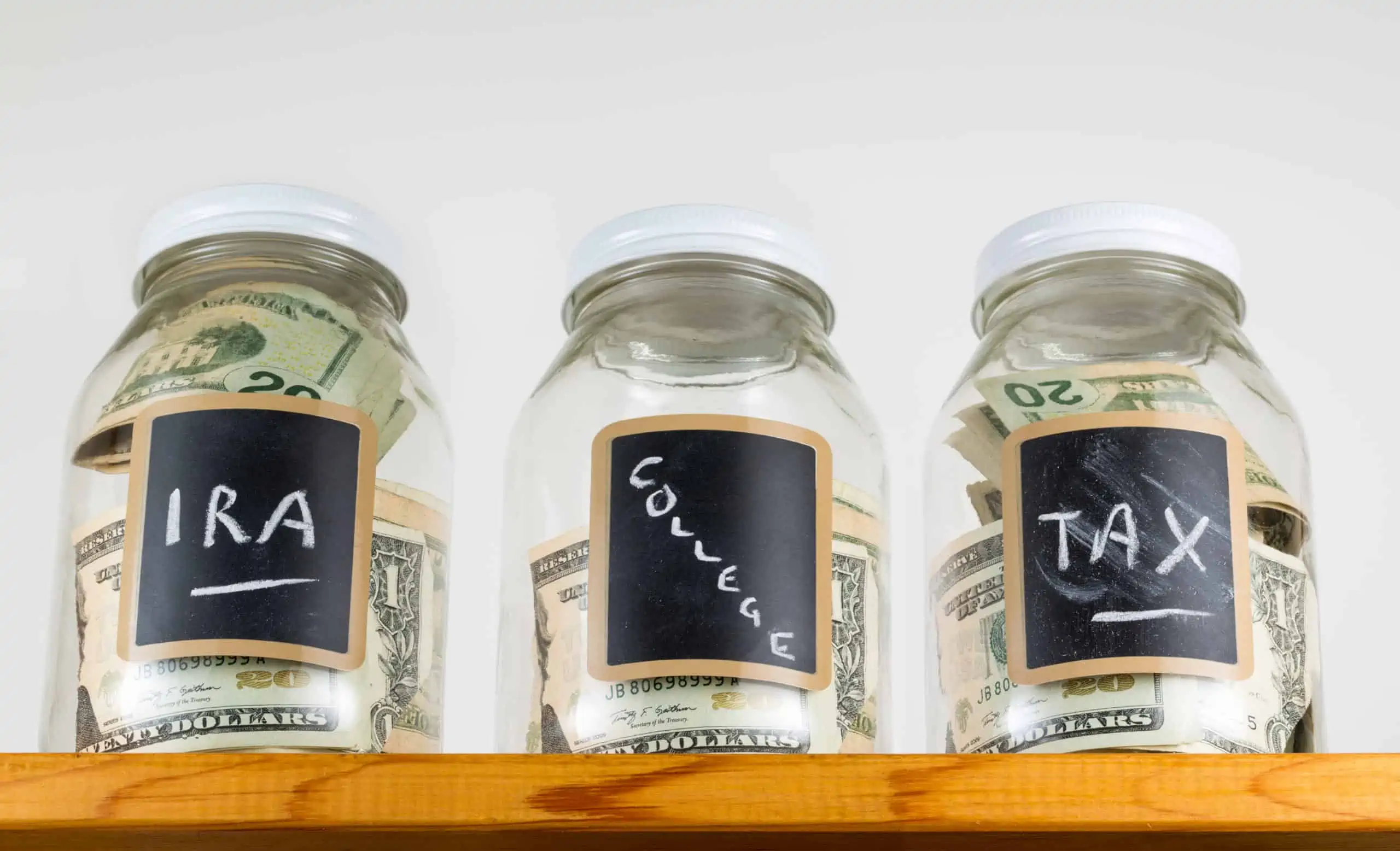 Three glass jars with chalk labels used for saving US dollar bills and notes for IRA, tax and college funds.