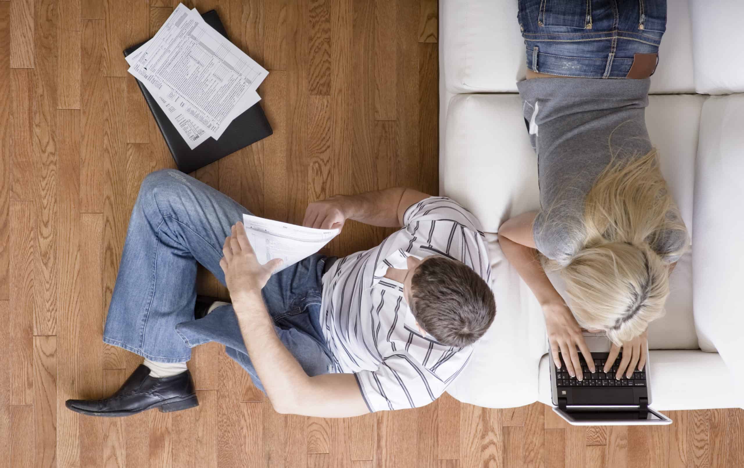 Caucasian woman laying on a white leather couch and a caucasian man sitting on a hardwood floor using tax apps to prepare taxes