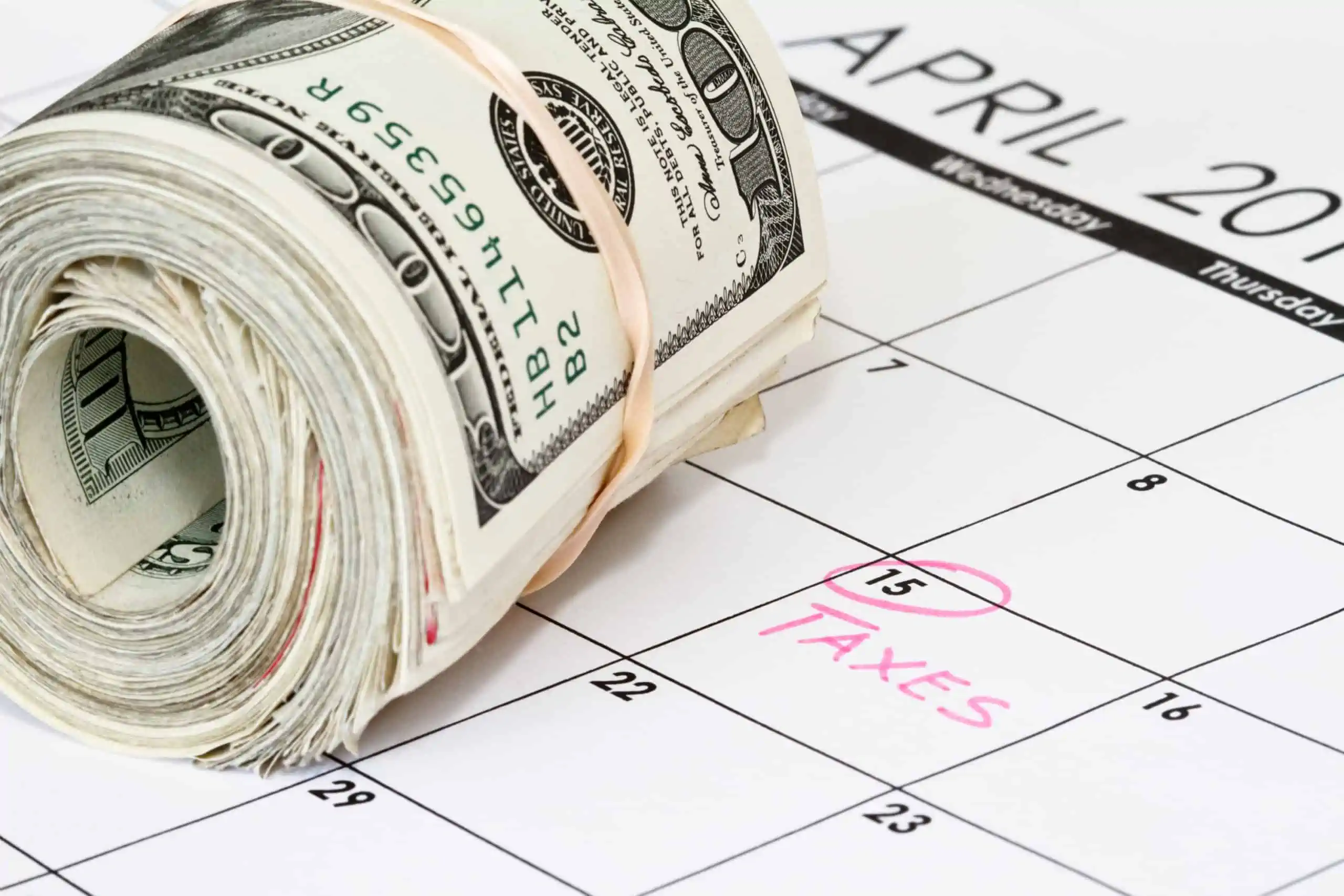 Tax filing deadline, April 15, circled on a calendar in pink with "taxes" written on it with a roll of $100 bills on top