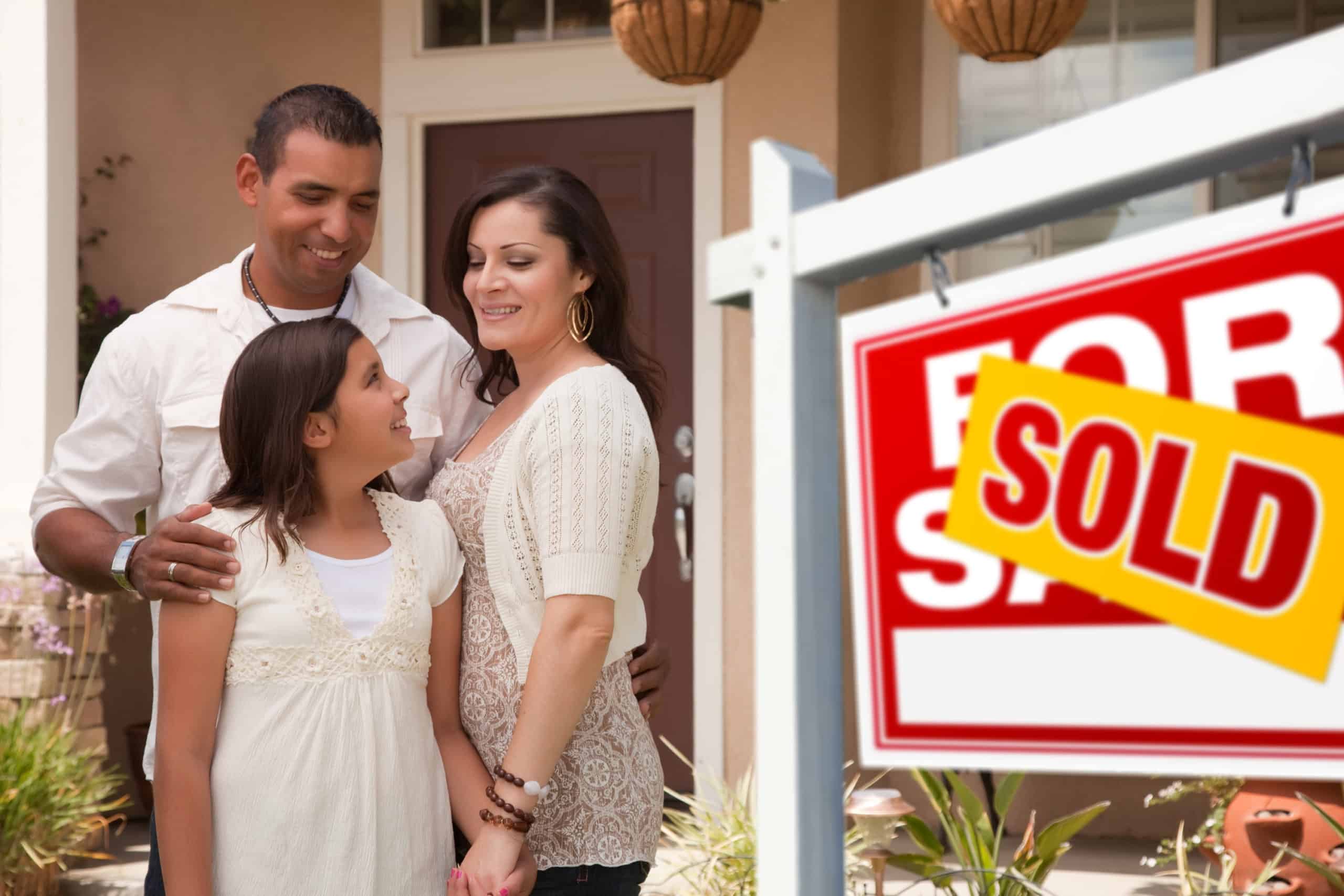 Hispanic mother, father, and daughter in front of their new home with for sale real estate sign.