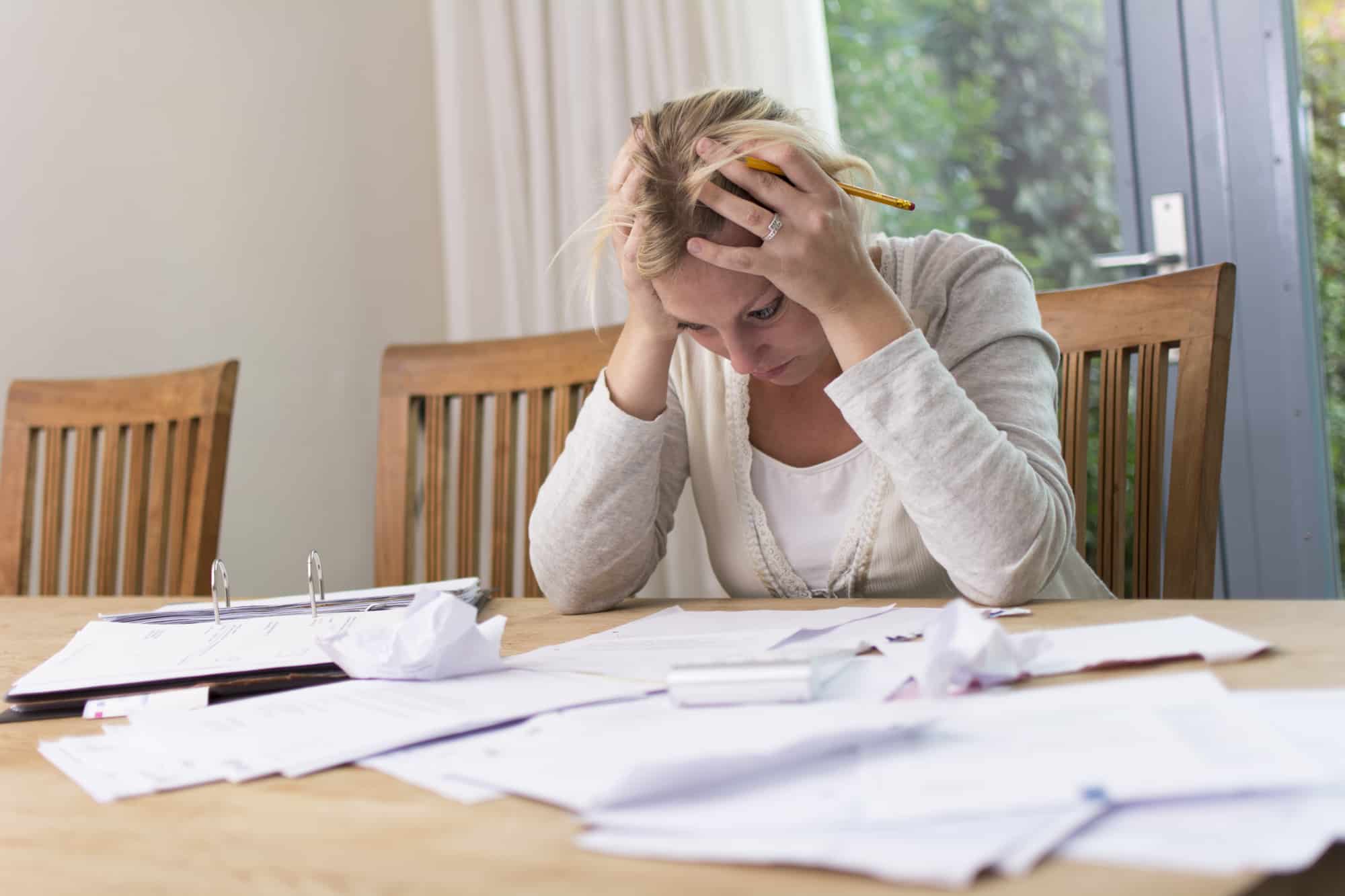 Older, blonde woman sitting at the dining room table holding her head in her hands with personal finances spread out.
